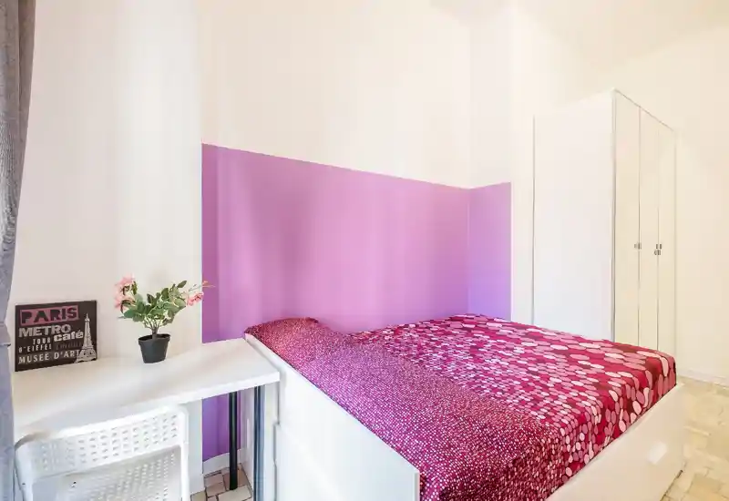 Camera singola luminosa con parete accentata rosa e copriletto fantasia, armadio bianco e una piccola scrivania con sedia — perfetta per un affitto compatto e accogliente.