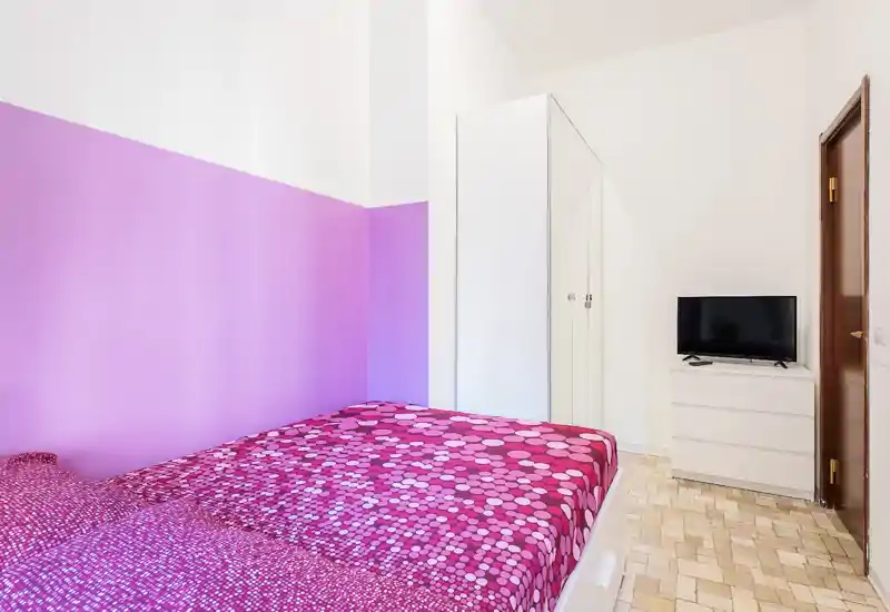 Camera da letto luminosa e moderna con parete accentuata rosa-viola, biancheria a motivi magenta, armadio e comò bianchi con TV: spazio accogliente e ben illuminato ideale per letto singolo o matrimoniale.