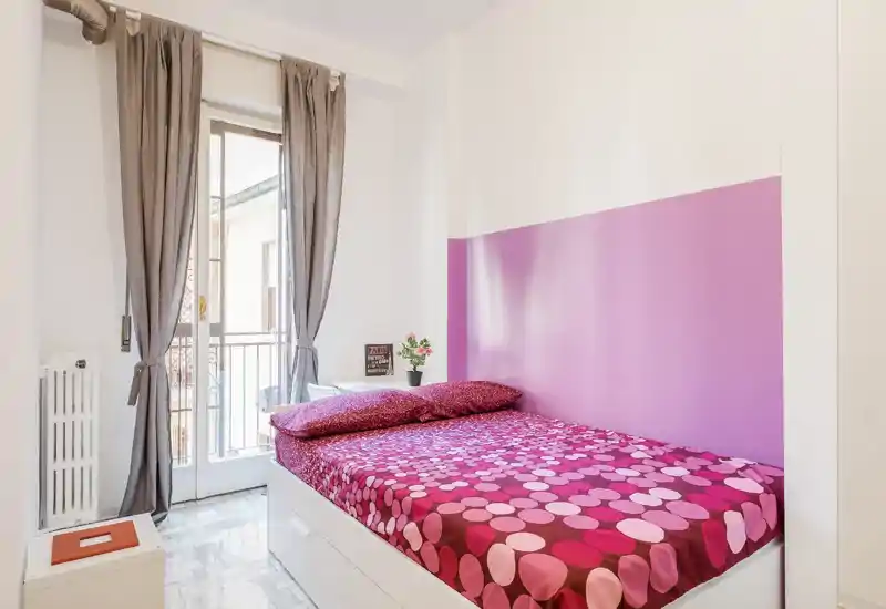 Camera da letto luminosa e compatta con letto singolo completo con biancheria rosa a motivi, parete con accento lavanda e porta balcone che fornisce molta luce naturale — ideale per un soggiorno cittadino accogliente.