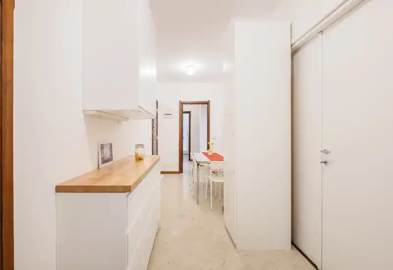 Corridoio cucina-pranzo stretto con mobili bianchi e piano in legno, piccolo tavolo da pranzo in fondo e stile minimal luminoso.