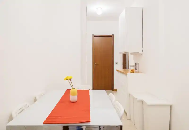 Zona pranzo minimal e luminosa con tavolo in vetro, runner rosso e sedie bianche; un piccolo vaso con fiori gialli aggiunge un accento caldo.
