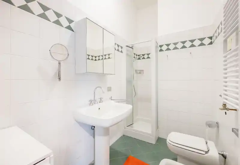 Bagno compatto e luminoso con lavabo a colonna, box doccia, bidet/wc e pensile specchiato. Piastrelle bianche pulite con decoro a rombi verdi e pavimento verde che donano un aspetto fresco.