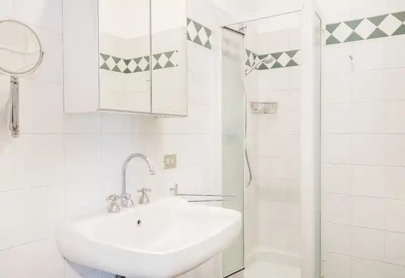 Bagno luminoso e pulito con lavabo bianco, mobiletto con specchio e cabina doccia compatta — piastrelle chiare e rubinetteria cromata perfette per un affitto breve.