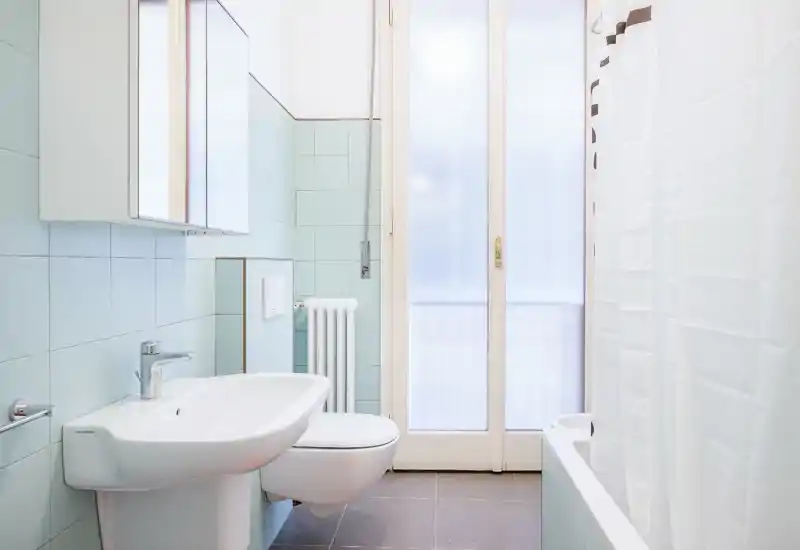 Bagno moderno e luminoso con wc sospeso, lavabo, vasca con tenda da doccia e porta a vetro satinato che lascia entrare luce naturale soffusa.