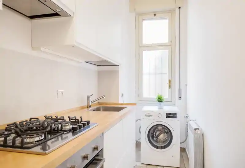 Cucina moderna compatta con piano cottura a gas integrato, lavello in acciaio e lavatrice frontale vicino alla finestra. Design luminoso e minimale con piano in legno e mobili bianchi ideale per appartamenti piccoli.