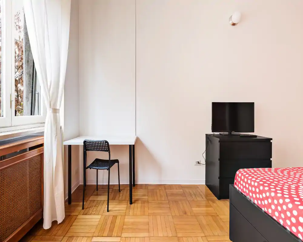 Angolo camera luminoso e minimal con copriletto a pois, una piccola scrivania vicino a una grande finestra e un comò con TV: linee pulite e luce naturale perfette per un annuncio.