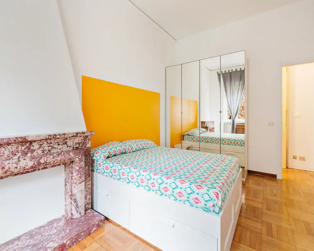 Camera da letto luminosa e moderna con un grande letto, biancheria dai colori vivaci, armadio con ante a specchio e un elegante camino in marmo. La luce naturale e le linee pulite creano un ambiente fresco e accogliente.