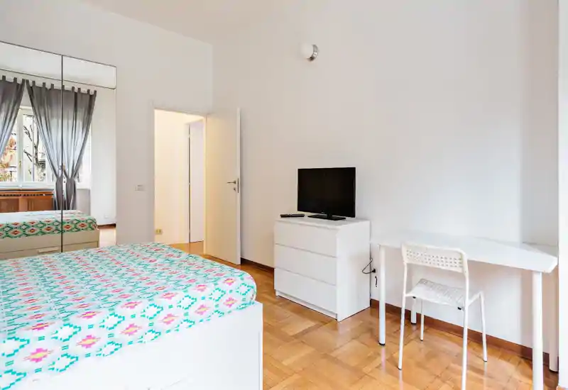 Camera da letto luminosa e minimal con copriletto colorato, comò bianco con TV e una piccola scrivania. Ampio armadio con specchi e finestra che garantiscono luce naturale e senso di spazio.