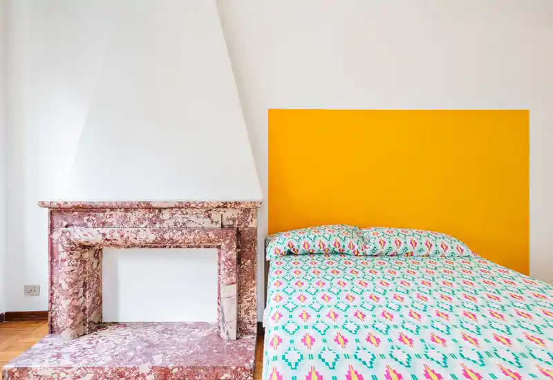 Camera da letto luminosa e di design con un letto matrimoniale con copriletto fantasia e un deciso pannello giallo come testiera accanto a un elegante camino in marmo rosa. Composizione pulita e colorata, ideale per annunci di affitto moderni.