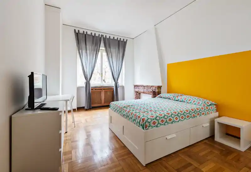 Camera da letto luminosa e ben arredata con letto matrimoniale, testiera colorata e luce naturale da una grande finestra — ideale per annunci.