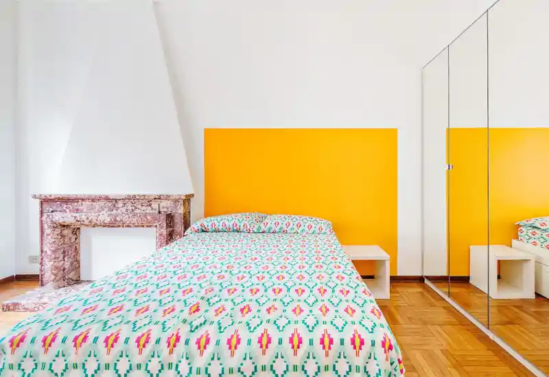 Camera da letto luminosa e moderna con letto completo in primo piano, biancheria dai motivi vivaci, parete accentata gialla e armadio con ante a specchio che ampliano la percezione dello spazio.