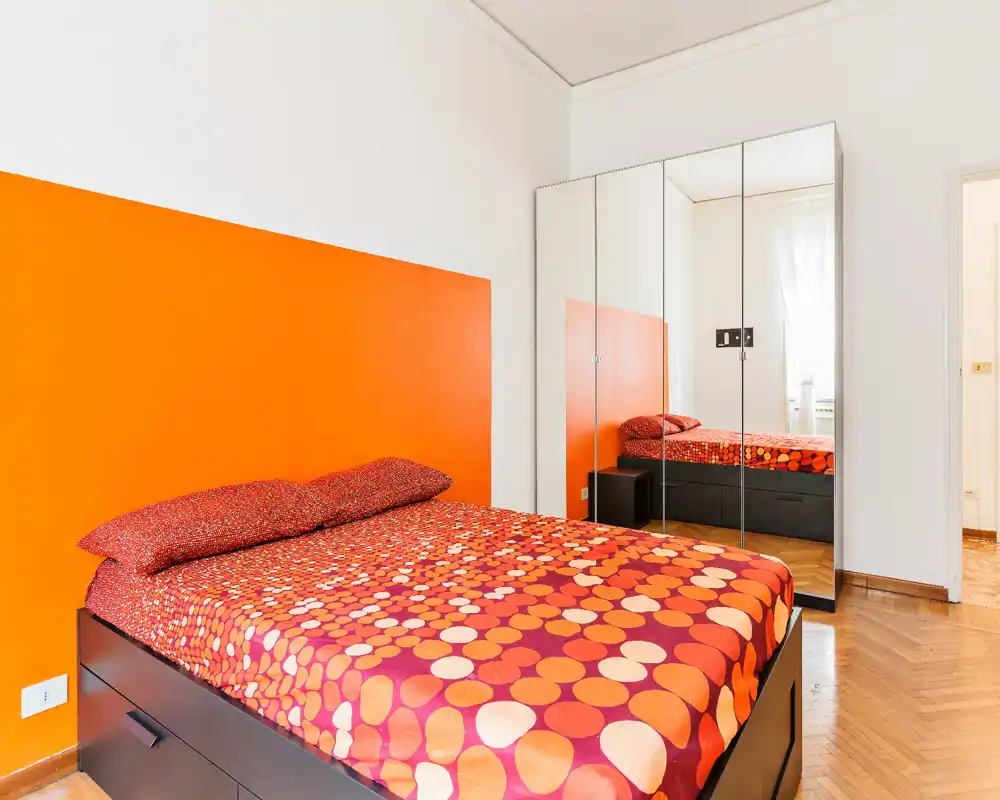 Camera da letto luminosa e moderna con testiera arancione vivace e biancheria a fantasia. Letto basso, pavimento in parquet e armadio con ante a specchio che amplifica la profondità.