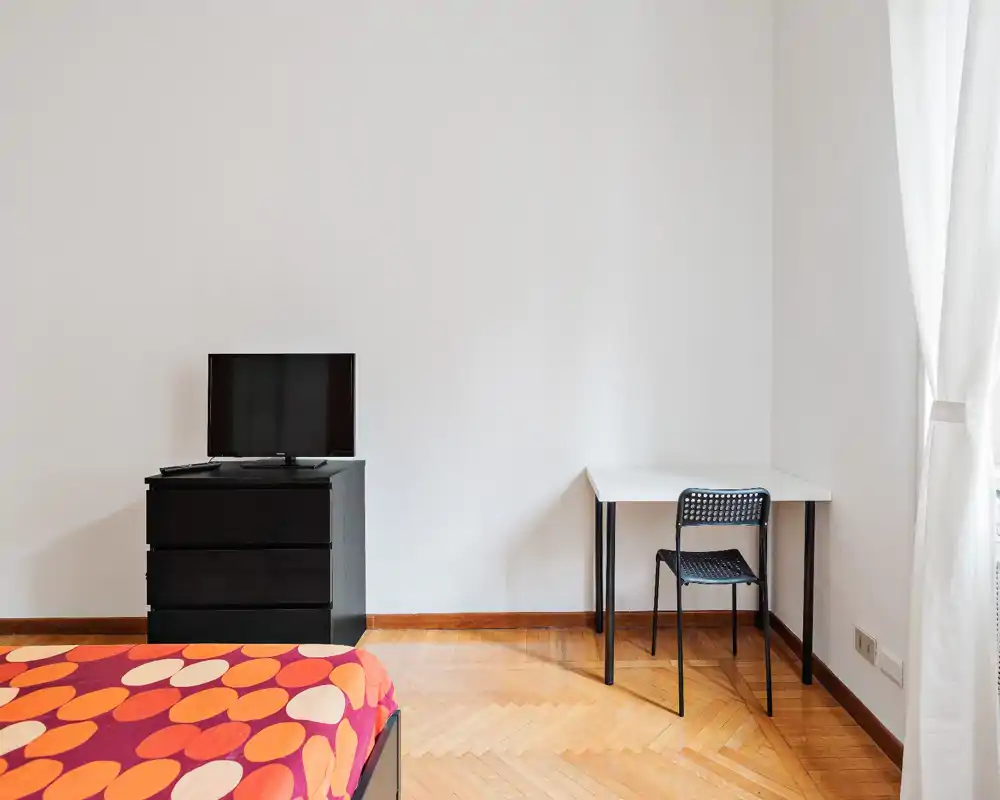 Camera da letto luminosa e minimale con copriletto colorato, TV su un comò nero e una scrivania semplice vicino alla finestra. La luce naturale e il pavimento in parquet creano un’atmosfera ariosa ideale per affitti brevi.