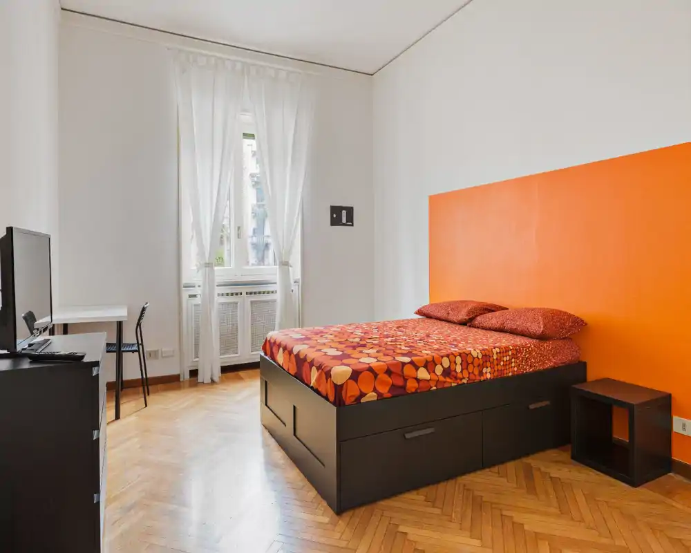 Camera da letto moderna e luminosa con un ampio letto matrimoniale, biancheria colorata e una parete arancione; la finestra alta e il pavimento in parquet donano calore.