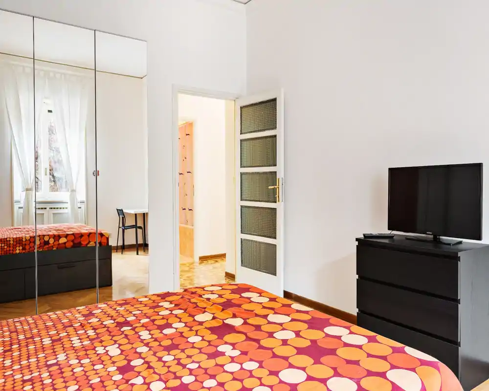 Camera da letto moderna e luminosa con copriletto arancione a motivi, armadio con ante a specchio e comò nero con TV. Ampia finestra che garantisce luce naturale e specchi che aumentano la percezione dello spazio.