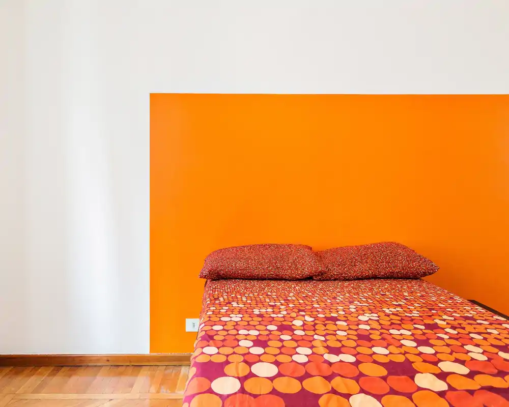 Camera da letto luminosa e moderna con una parete arancione e un letto matrimoniale con copripiumino a fantasia. Linee pulite, pavimento in legno caldo e arredamento essenziale creano un ambiente accogliente e contemporaneo.
