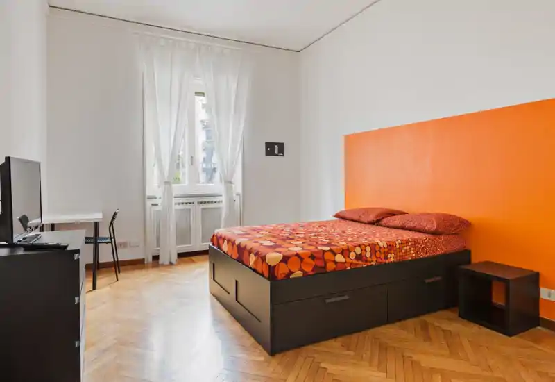 Camera da letto moderna e luminosa con un ampio letto matrimoniale, biancheria colorata e una parete arancione; la finestra alta e il pavimento in parquet donano calore.