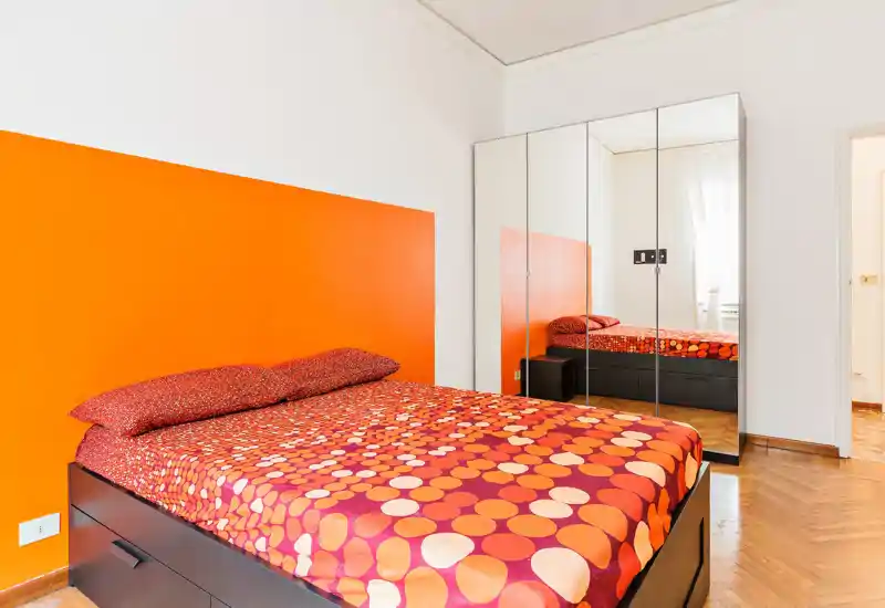 Camera da letto luminosa e moderna con letto grande con biancheria arancione/rossa a motivi, testiera arancione vivace e armadio con ante a specchio che ampliano lo spazio e la luce naturale.