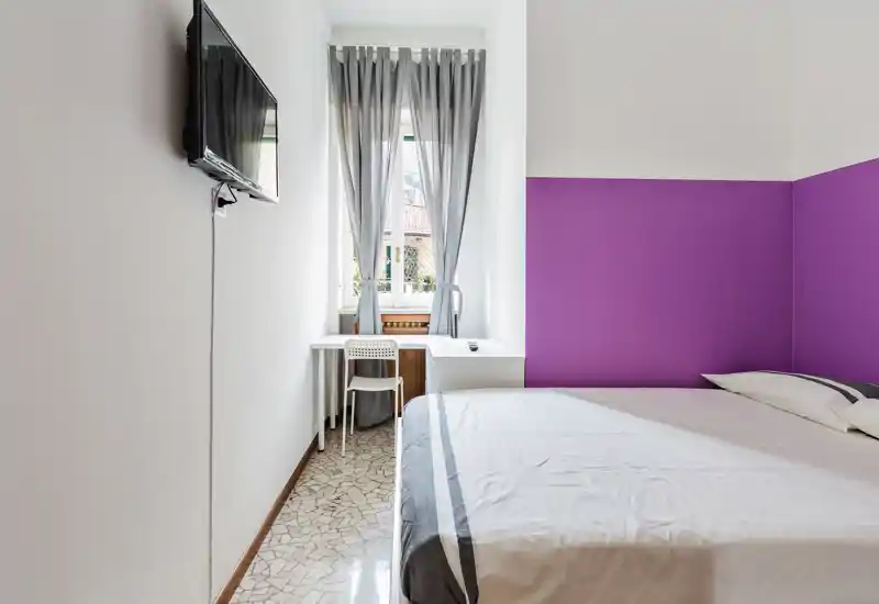 Camera da letto luminosa e compatta con letto matrimoniale, parete viola come elemento d'accento e una piccola scrivania davanti alla finestra con luce naturale.