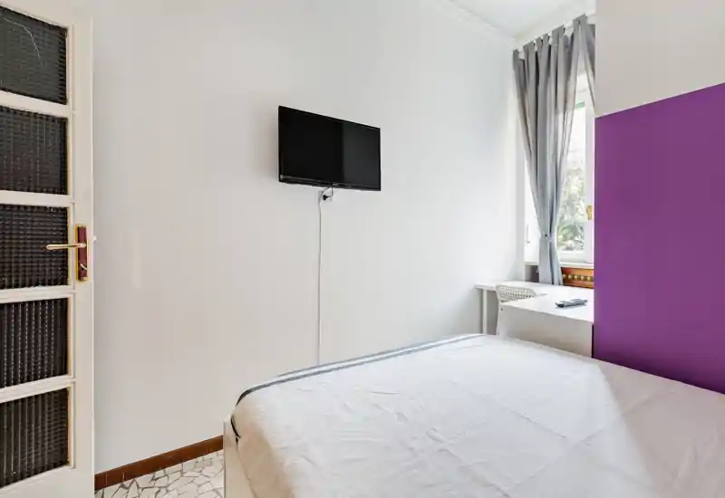 Camera da letto luminosa e arredata in modo semplice con letto in primo piano, TV a parete e una scrivania vicino alla finestra con luce naturale.