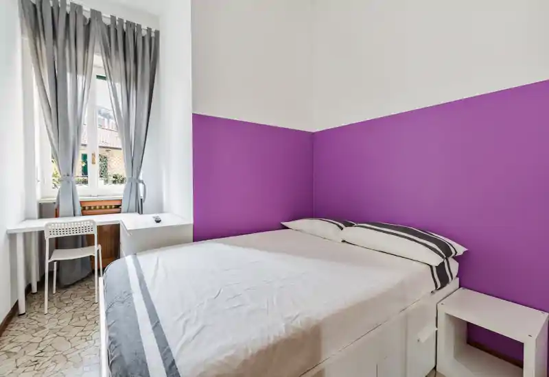 Camera da letto compatta e luminosa con letto matrimoniale contro una parete viola a mezza altezza, semplice comodino bianco e una piccola scrivania vicino alla finestra che lascia entrare luce naturale.