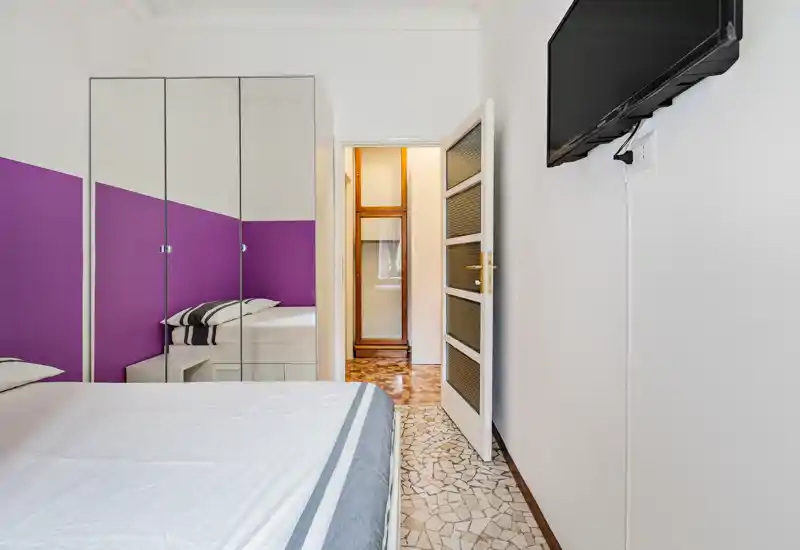 Camera da letto luminosa e moderna con letto matrimoniale, parete viola a contrasto e armadio con ante a specchio che amplia visivamente lo spazio. Linee pulite e pavimento a mosaico donano un aspetto fresco e ordinato, perfetto per affitti brevi.