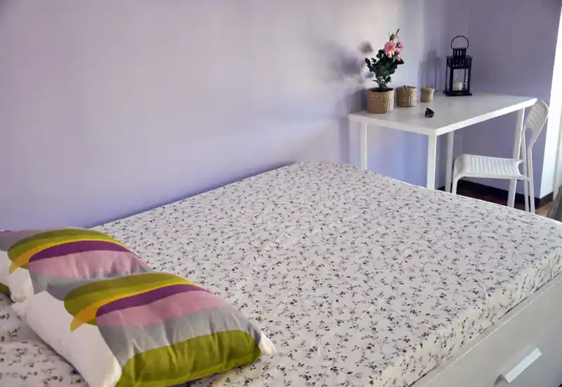 Camera da letto luminosa e accogliente con un ampio letto matrimoniale con lenzuola floreali e un cuscino colorato; piccolo scrittoio con pianta decorativa e lanterna vicino alla finestra.