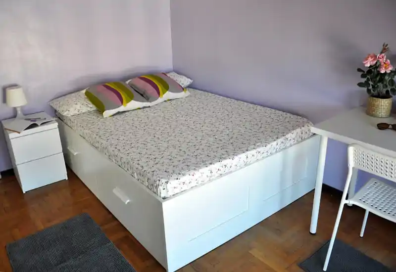 Camera da letto luminosa e ordinata con un ampio letto matrimoniale, biancheria floreale, comodino con lampada e una piccola scrivania con sedia e pianta — perfetta per una persona o coppia.