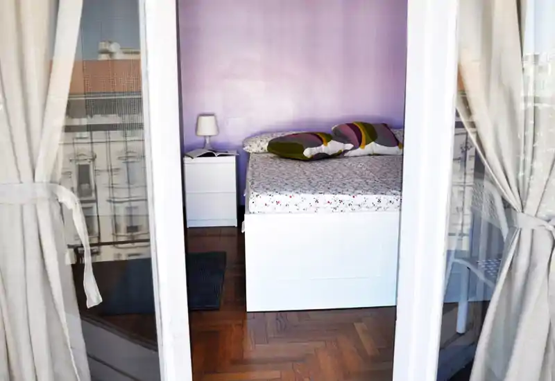 Camera da letto luminosa e accogliente vista dalla porta del balcone: letto bianco con biancheria a fantasia, cuscini decorativi, comodino con lampada e pavimento in parquet.
