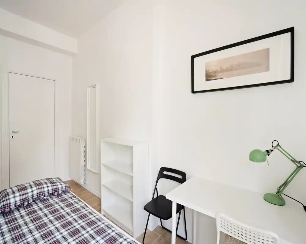 Bright, minimal bedroom with a single bed, white shelving and a simple desk with a green lamp — ideal for a student or short-term rental.