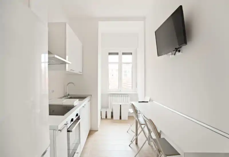 Cocina luminosa y minimalista con armarios blancos, electrodomésticos integrados y una encimera estrecha para comer junto a la pared; una ventana aporta luz natural y una TV en la pared.