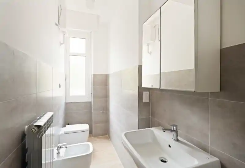 Modern, bright bathroom with wall-mounted mirror cabinet, sink, bidet and toilet; neutral gray tiles and a tall frosted window provide soft natural light.