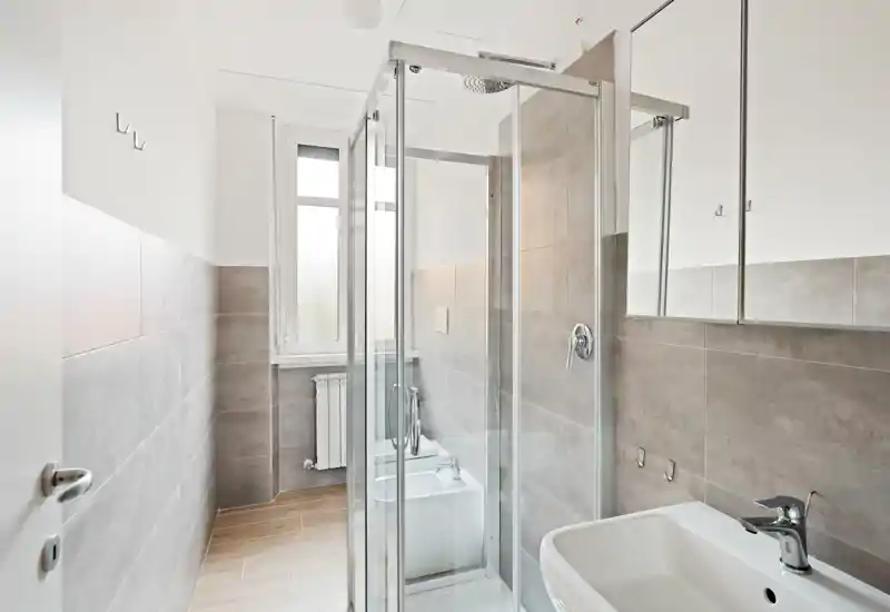 Modern, bright bathroom with a glass shower enclosure, wall-mounted bidet/toilet fixtures, sink with chrome faucet and mirrored cabinet — clean neutral tiles and natural light from the window.