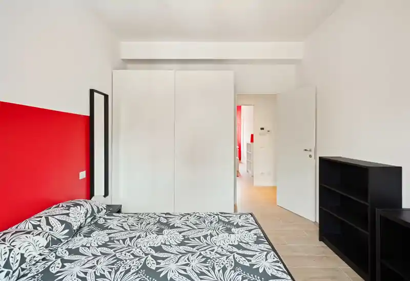 Dormitorio moderno y luminoso con cama doble estampada, pared roja de acento y mobiliario minimalista (armario y estantería). La habitación está ordenada, bien iluminada y preparada para fotos de alquiler.