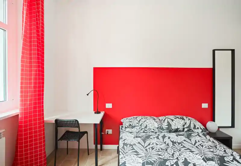 Dormitorio luminoso y moderno con cama doble estampada, pared roja de acento y un pequeño escritorio junto a la ventana — ideal para una estancia compacta y con estilo.