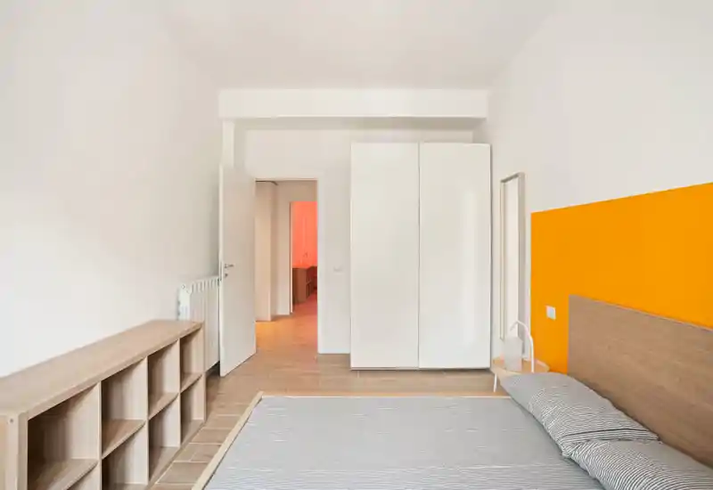 Camera da letto luminosa e minimal con letto matrimoniale contro una parete arancione, testiera in legno, armadio e scaffalatura aperta. Linee pulite e luce naturale ideali per annunci moderni.