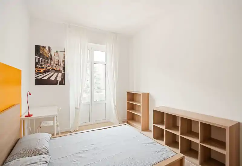 Camera da letto luminosa e minimal con letto matrimoniale, scaffalature in legno semplici e una scrivania vicino alla finestra che illumina la stanza con luce naturale.