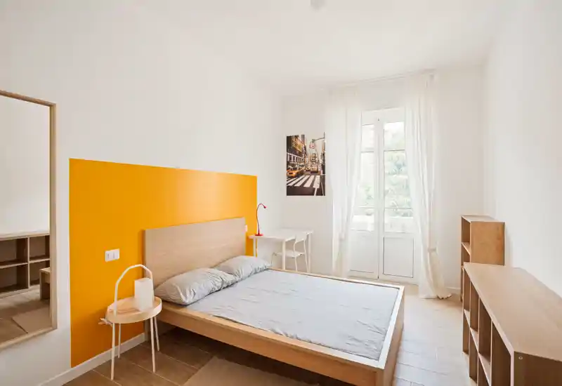 Camera da letto luminosa e minimal con letto matrimoniale in legno, pannello arancione sul muro, comodino, piccolo scrittoio vicino alla finestra e scaffalatura — ariosa e ben illuminata.