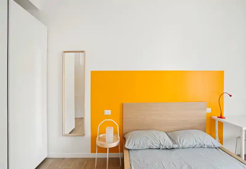 Camera da letto luminosa e minimal con letto in legno e pannello giallo dietro la testata, biancheria a righe, piccolo comodino e specchio sottile — stile moderno e accogliente, ottimo per annunci.