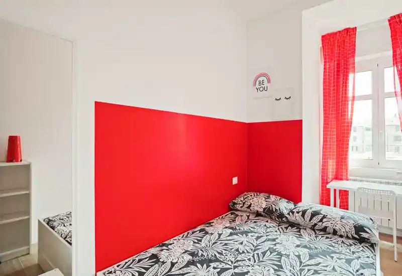Dormitorio compacto y luminoso con cama doble junto a una pared roja intensa, ropa de cama estampada y una ventana que aporta mucha luz natural.