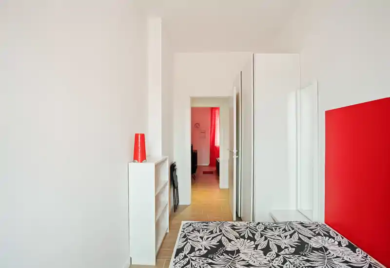 Dormitorio minimalista con colchón estampado, cabecero rojo y estantería blanca. Espacio luminoso y limpio, con un pasillo que conduce a otra habitación con cortinas rojas.