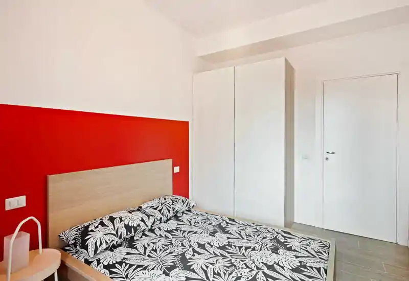 Dormitorio moderno y luminoso con cabecero de madera, ropa de cama estampada y armario blanco; la pared roja aporta un toque cálido y atrevido.