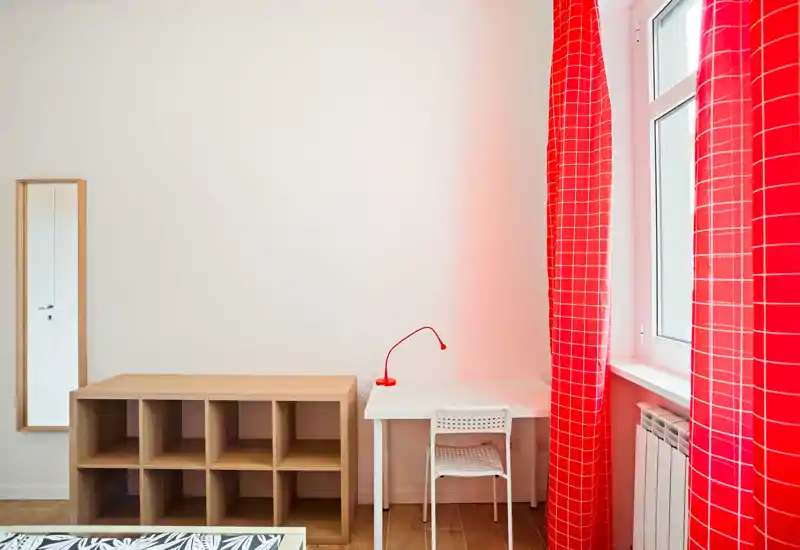 Rincón de dormitorio minimalista y luminoso con escritorio blanco, silla y lámpara de lectura roja junto a la ventana con cortinas rojas; estantería de madera y un destello de la cama en el borde de la foto completan el ambiente moderno.