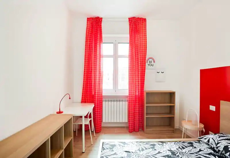Dormitorio moderno y luminoso con llamativas cortinas rojas y colcha estampada; estanterías de madera y un pequeño escritorio crean un espacio funcional y ordenado, ideal para alquileres temporales.