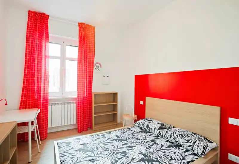 Dormitorio moderno y luminoso con cama doble, ropa de cama estampada, cabecero de madera y pared de acento roja. La luz natural y las cortinas rojas coordinadas aportan una atmósfera fresca y acogedora, perfecta para un anuncio.