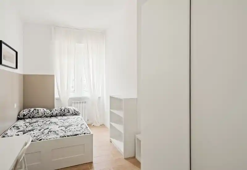 Dormitorio luminoso y minimalista con cama doble con ropa de cama estampada, ventana alta con cortinas translúcidas y muebles blancos de almacenamiento — limpio y muy luminoso.