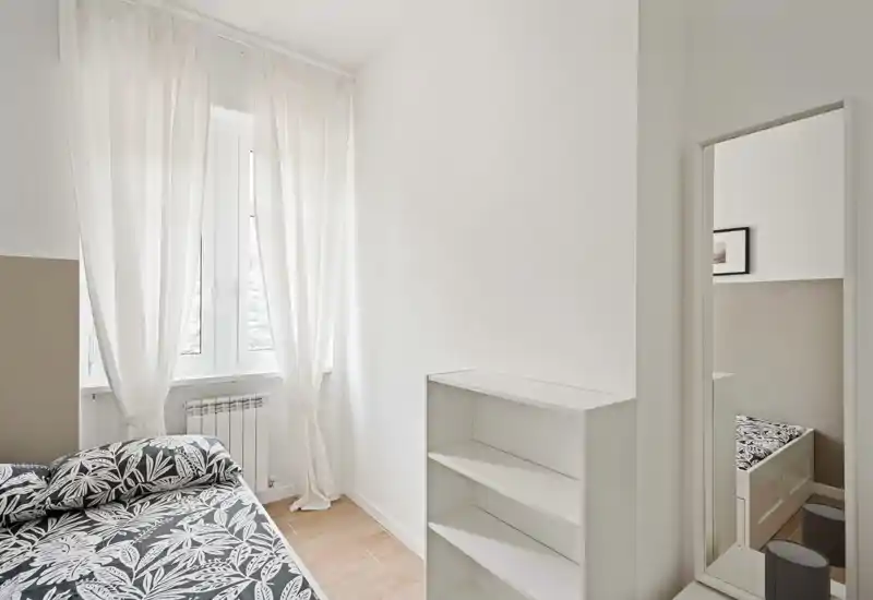 Dormitorio minimalista y luminoso con ropa de cama estampada, una gran ventana con cortinas ligeras y estanterías sencillas. El espacio es aireado y ordenado, ideal para alquileres de corto plazo o una habitación de invitados.