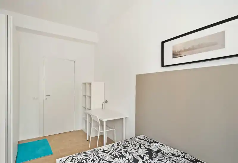 Dormitorio luminoso y minimalista con un edredón estampado, un pequeño escritorio y estantería junto a la puerta — estilo moderno y tonos neutros.