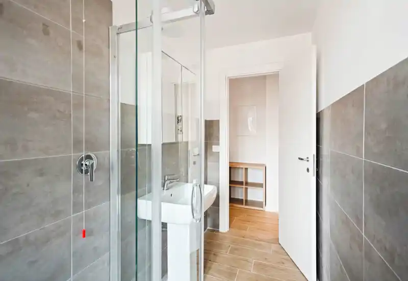 Baño moderno con ducha de cristal, lavabo y paredes con azulejos grises; luminoso, limpio y de diseño minimalista, ideal para anuncios de alquiler.