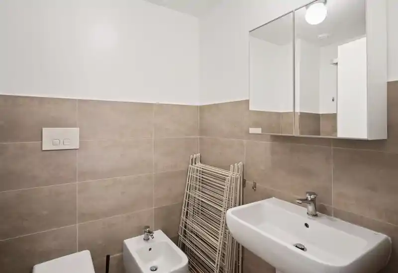 Baño moderno y limpio con armario con espejo, lavabo, bidé y WC parcialmente visibles; azulejos beige neutro e iluminación clara y uniforme.